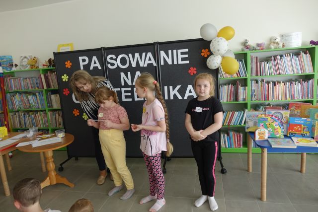 trójka dzieci stoi z kobietą, w tle regały z książkami