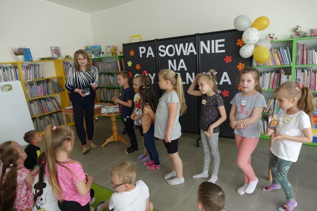 6 dzieci i kobieta stoją przed dziećmi siedzącymi