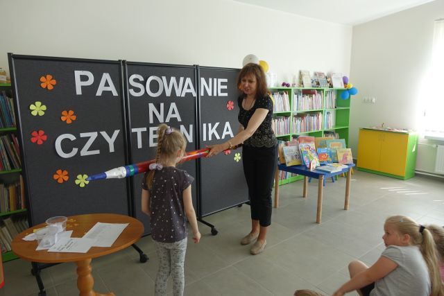 kobieta ubrana na czarno pasuje ogromnym ołówkiem dziewczynkę na czytelniczkę