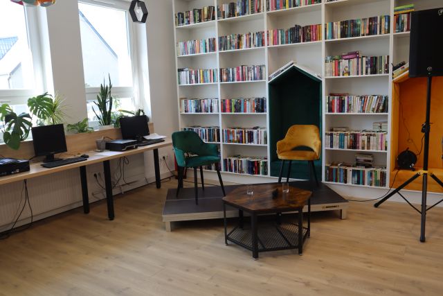 dwa krzesła zielone i żółte ustawione na podwyższeniu, w tle białe regały z książkami