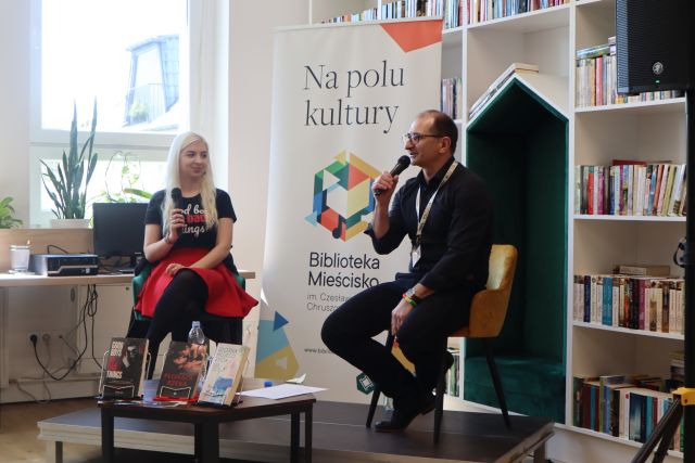 kobieta w blond włosach i mężczyzna ubrany na czarno siedzą na kolorowych krzesłach na podwyższeniu