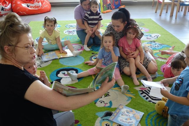 dzieci i opiekunowie siedzą na kolorowym dywanie i czytają książki