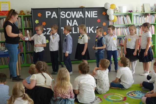 grupka przedszkaloków stoi na środku przed siedzącymi na kolorowym dywanie dziećmi