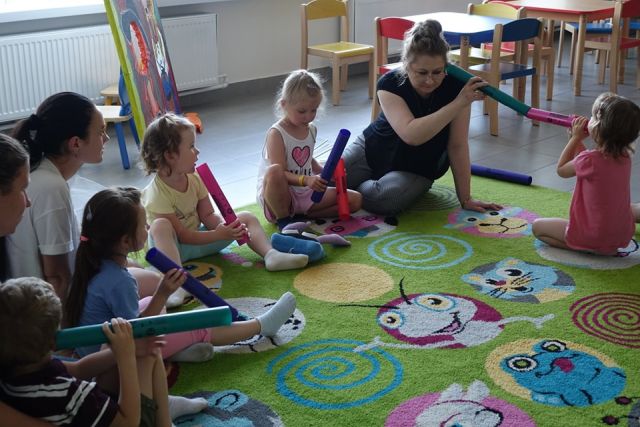 dzieci i rodzice siedzą na kolorowym dywanie i bzwią się w głuchy telefon wykorzystując do tego bum bum rurek