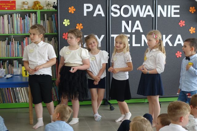 grupka przedszkaloków stoi na środku przed siedzącymi na kolorowym dywanie dziećmi