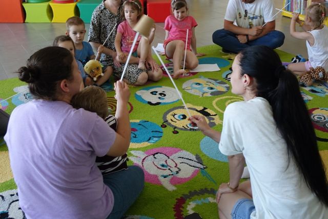 dzieci i rodzice siedzą na kolorowym dywanie i przy pomocy długich patyczkó podają sobie papierowe kubeczki