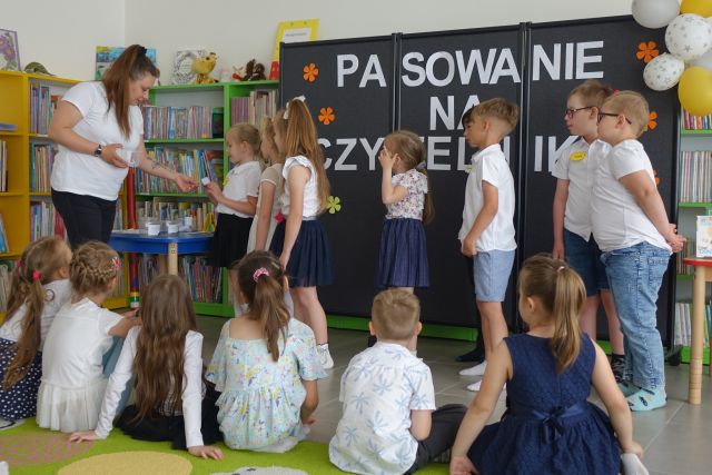 grupka przedszkaloków stoi na środku przed siedzącymi na kolorowym dywanie dziećmi
