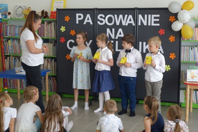 grupka przedszkaloków stoi na środku przed siedzącymi na kolorowym dywanie dziećmi