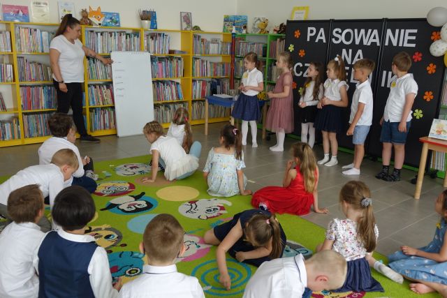 grupka przedszkaloków stoi na środku przed siedzącymi na kolorowym dywanie dziećmi