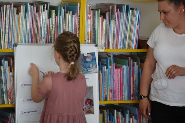 dziewczynka przypina do tablicy magnetycznej obrazki z bajek, w tle regały z książkami