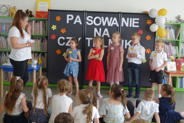 grupka przedszkaloków stoi na środku przed siedzącymi na kolorowym dywanie dziećmi