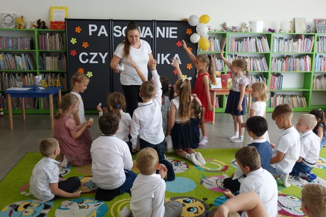 dzieci siedzą na dywanie i podnoszą do góry paluszki, przed nimi stoi kobieta, w tle regały z książkami