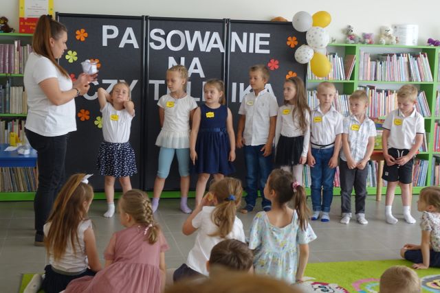 grupka przedszkaloków stoi na środku przed siedzącymi na kolorowym dywanie dziećmi