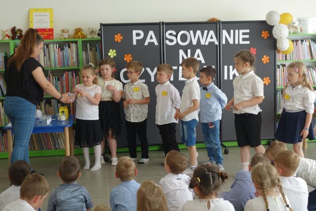 grupka przedszkaloków stoi na środku przed siedzącymi na kolorowym dywanie dziećmi