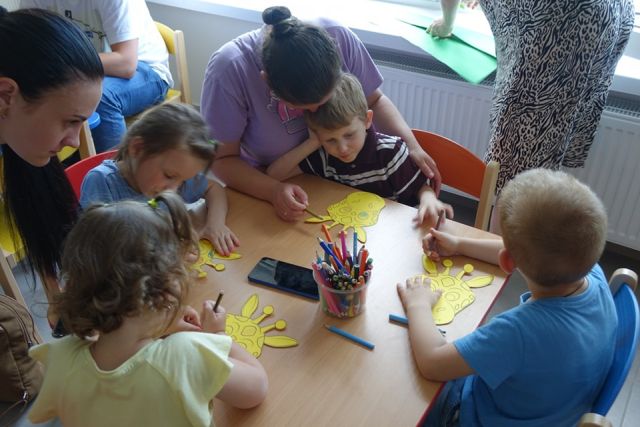 dzieci i rodzice siedzą przy stolikach i wykonują żyrafy z papieru
