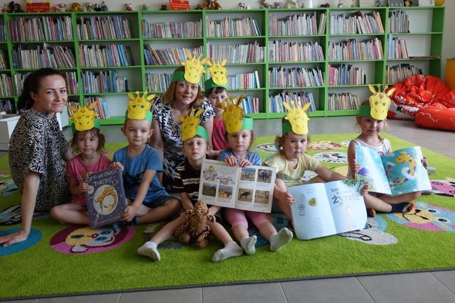 dzieci z rodzicami pozują do zdjęcia w maskach żyrafy trzymając w ręce książki, w tle regały z książkami