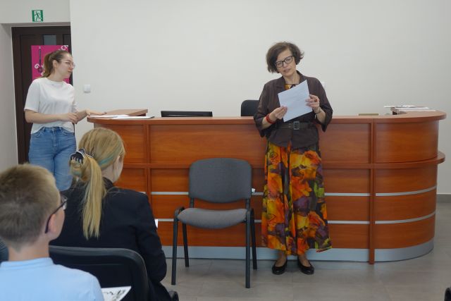 kobieta stoi przed uczestnika,i konkursu i czyta, za nią biurko