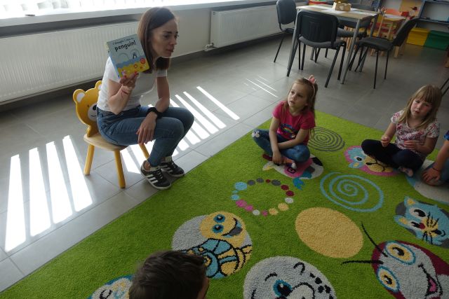 kobieta siedzi na krzesełku przed dziećmi siedzącymi na kolorowym dywanie i pokazuje książkę