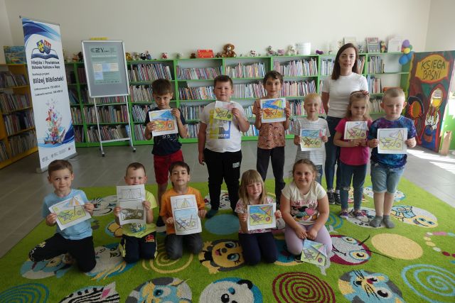 dzieci i kobieta pozują do zdjęcia i pokazują wykonane przez siebie ilustracje, w tle regały z książkami