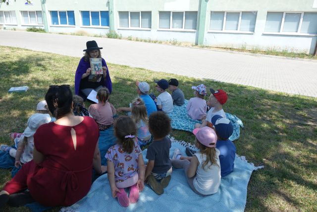 dzieci siedzą na kocykach na trawie i patrzą na kobietę przebraną za kota w butach trzymającą w dłoniach książkę