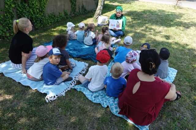 Kobieta przebrana za żabę czyta dzieciom siedzącym na trawie książkę
