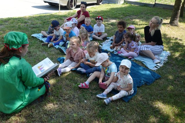 kobieta przebrana za żabę czyta dzieciom siedzącym na trawie książkę