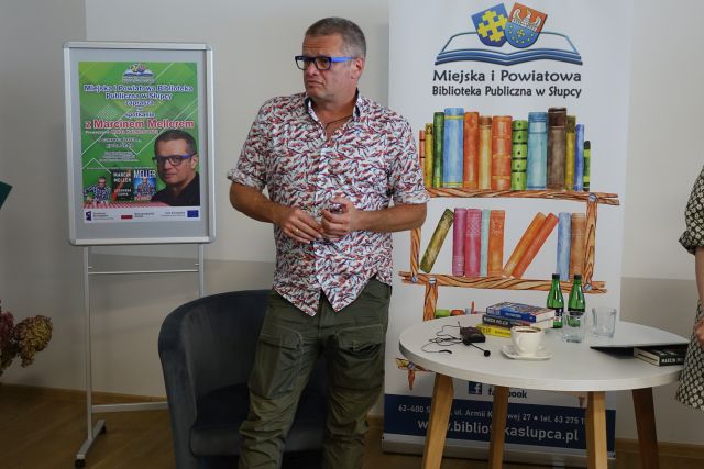 mężczyzna stojący, za nim siwy fotel i baner biblioteki