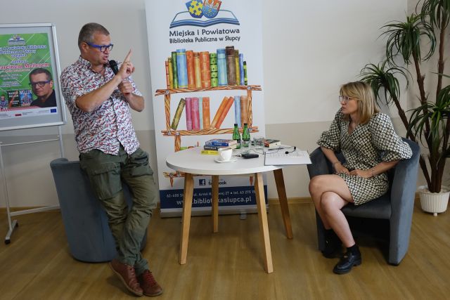 kobieta siedzi z prawej strony na siwym fotelu, z lewej stoi oparty o fotel mężczyzna, pomiędzy nimi stolik, w tle baner biblioteki
