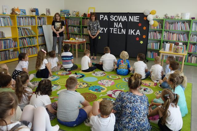 dzieci siedzą na kolorowym dywanie, przed nimi stoją dwie kobiety