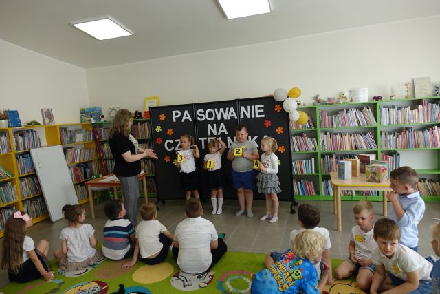 przedszkolaki siedzą na kolorowym dywanie, przed nimi stoją inne dziecii kobieta