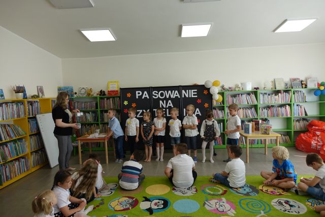 przedszkolaki siedzą na kolorowym dywanie, przed nimi stoją inne dziecii kobieta