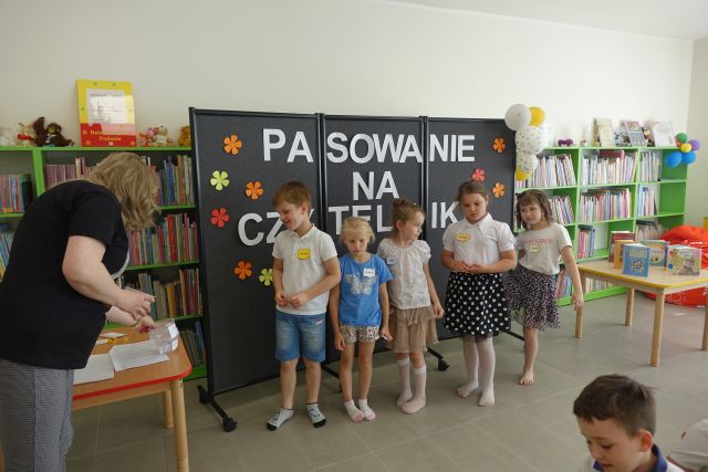 dzieci stoją na tle czarnej tablicy z napisem Pasowanie na Czytelnika