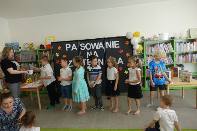 dzieci stoją na tle czarnej tablicy z napisem Pasowanie na Czytelnika