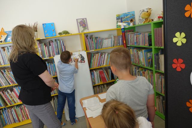chłopiec przypina do tablice ilustracje z bajek, w tle regały z książkami