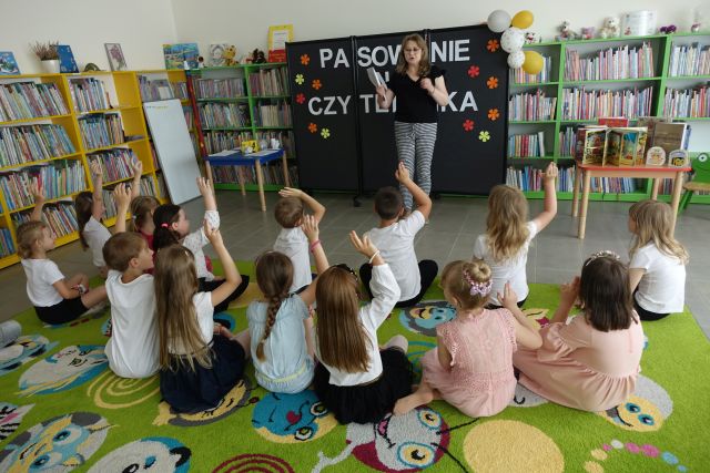 kobieta stoi przed dziećmi siedzącymi na kolorowym dywanie i zgłaszającymi się do odpowiedzi