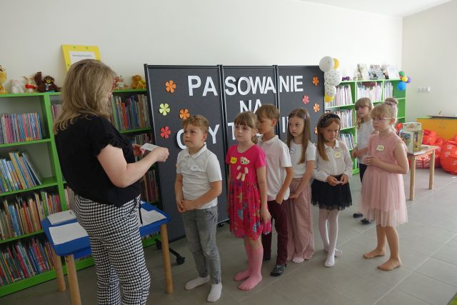 dzieci stoją na tle czarnej tablicy, przed nimi kobieta czyta z kartki