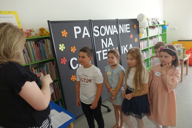 dzieci stoją na tle czarnej tablicy, przed nimi kobieta czyta z kartki