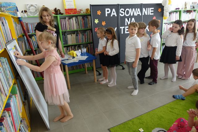 dzieci stoją na tle czarnej tablicy, dziewczynka przypina do białej tablicy ilustrację, obok stoi kobieta