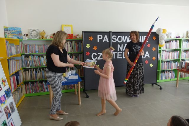 dziewczynka odbiera nagrodę od kobiety, z prawej strony stoi inna kobieta z ogromnym ołówkiem