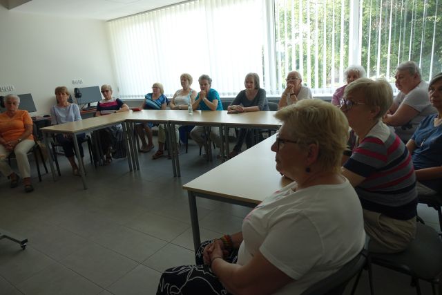 seniorki siedzą na krzesłąch przy stołąch ustawionych w podkowę w sali komputerowej