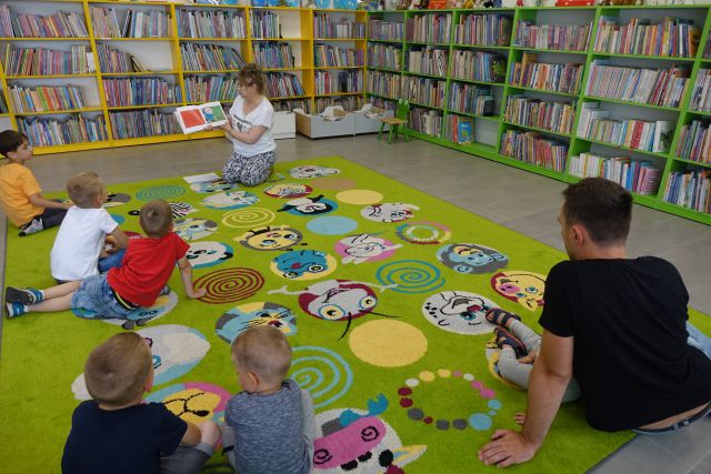 dzieci, rodzice, bibliotekarka siedzą na kolorowym dywanie i słuchają opowiadania z książki., w tle regały z książkami