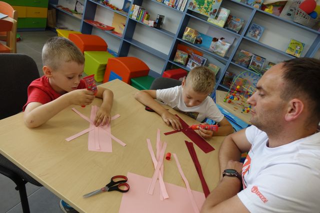 dwóch chłopców i mężczyzna siedzą przy stoliku i sklejają paski