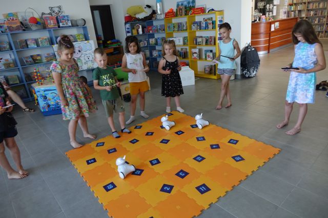 dzieci sterują robotami przy użyciu tabletów, w tle regały z książkami