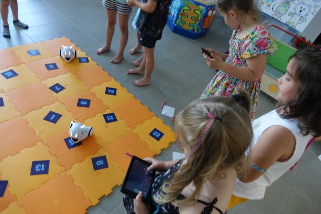 dzieci sterują przy użyciu tabletów robotami poruszającymi się po pomarańczowej macie