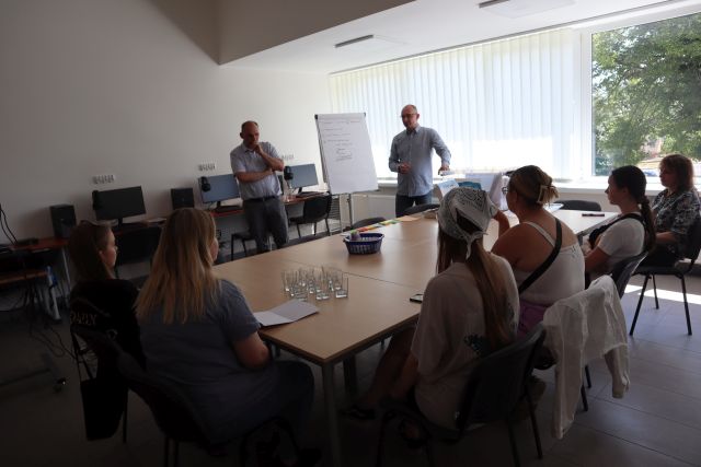 5 uczestniczek warsztatów siedzi przy stole i słyuch tłumaczeń dwóch mężczyzn. W tle sala komputerowa