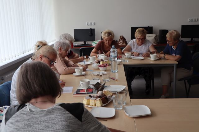 seniorki siedzą przy stołąch z ciastem i wodą i rozwiązują zadania
