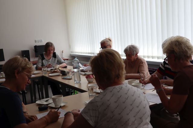 seniorki siedzą przy stołąch z ciastem i wodą i rozwiązują zadania