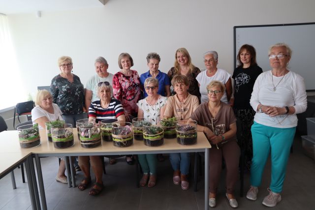 na stólach stoją stworzone przez seniorki ogrody w słoikach, seniorki pozują do zdjęcia siedząc i stojąc za stołami