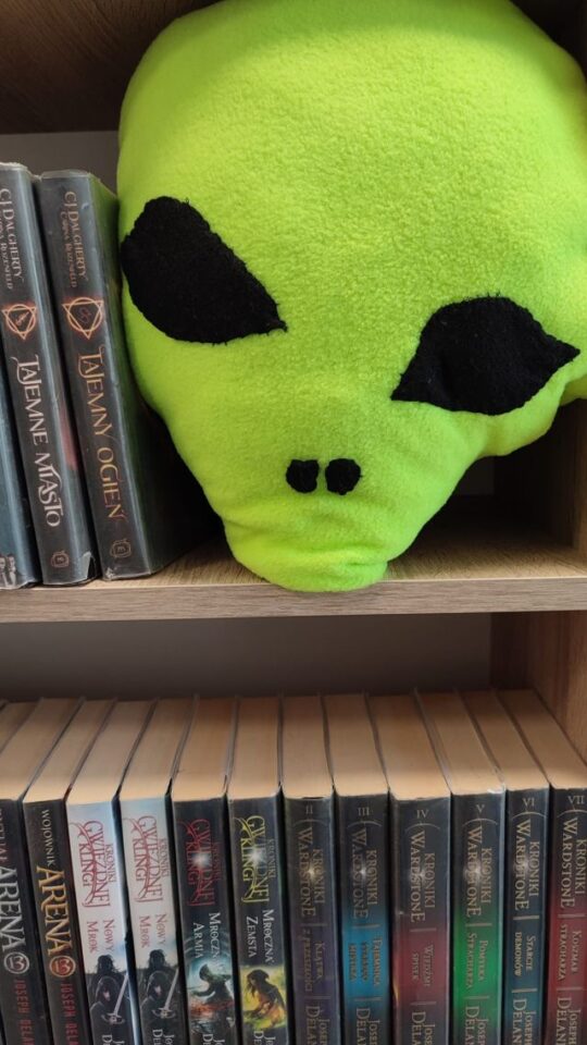maskotka zielonego ufoludka ułożona na ksiązkach