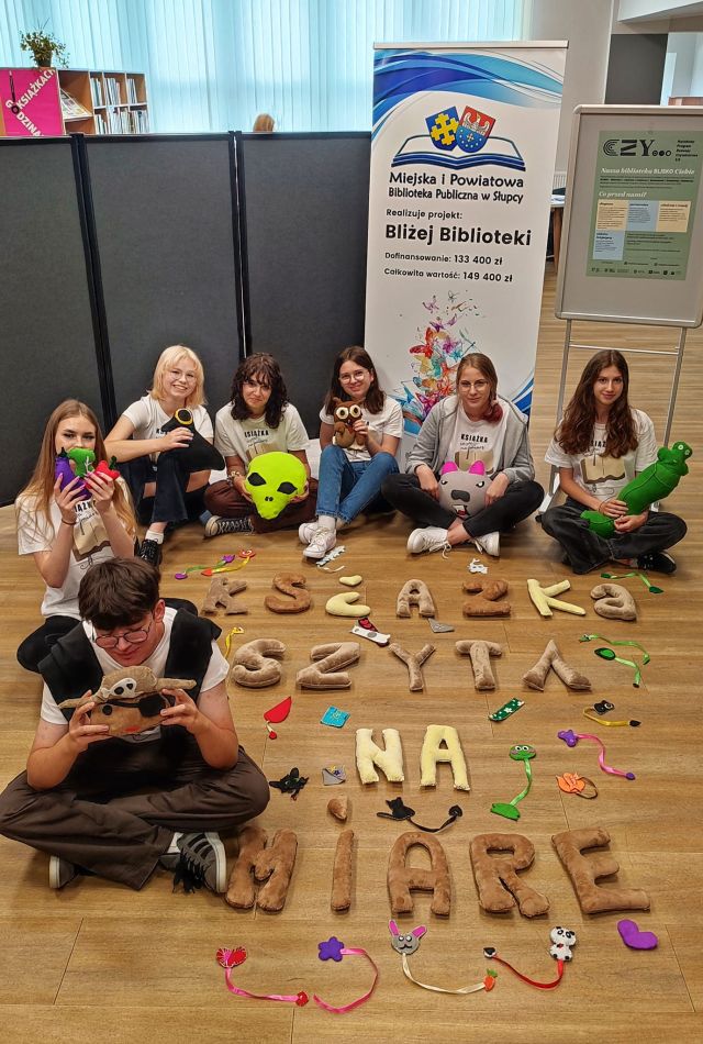 młodzież siedząca na podłodze pozuje do zdjęcia z uszytymi przez siebie maskotkami, napisem książka szyta na miarę i zakładkami do książek
